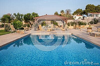 Reflective Blue Pool With A Wooden Pergola And Lounging Chairs In The Background Symbolizing