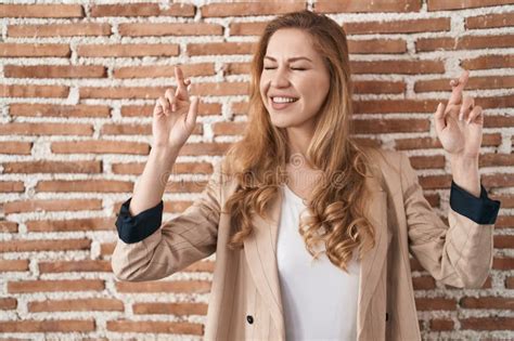 Beautiful Blonde Woman Standing Over Bricks Wall Gesturing Finger Crossed Smiling With Hope And