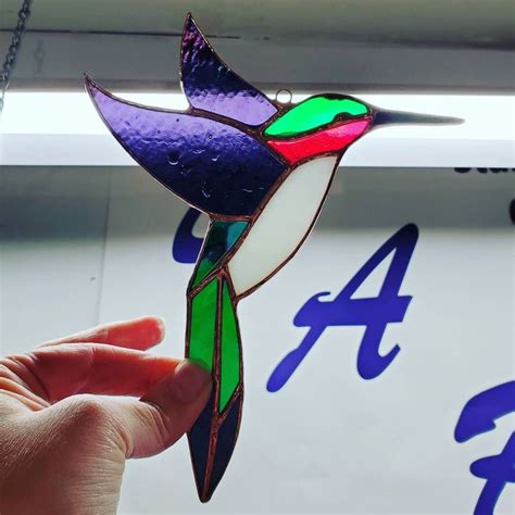 A Hand Holding A Stained Glass Hummingbird Ornament In Front Of A Sign