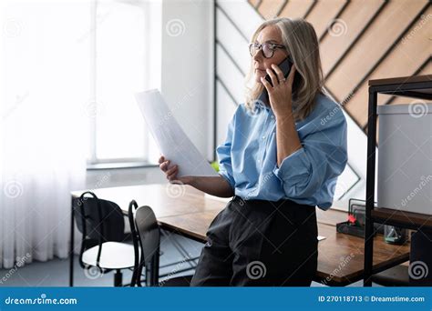 Mature Adult Caucasian Business Woman Talking On Mobile Phone While Looking At Documents While
