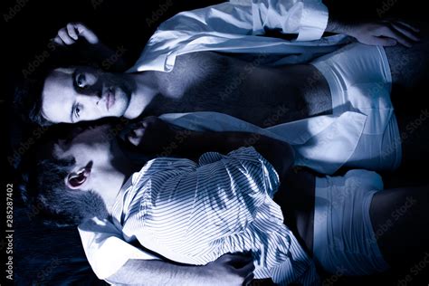 Gay Male Couple Lying In Bed With One Of Them Looking At Camera As Other Is Holding Him Stock