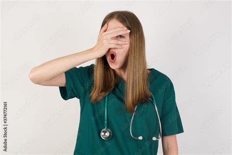 Female doctor wearing a green scrubs and stethoscope peeking in shock covering face and eyes ...