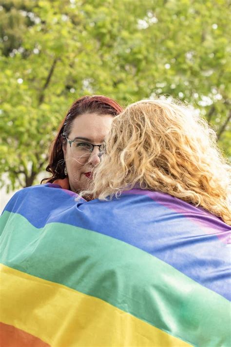 Lesbian Couple Embracing Under LGBT Rainbow Flag One Of Them Is Looking At Camera Concept Of