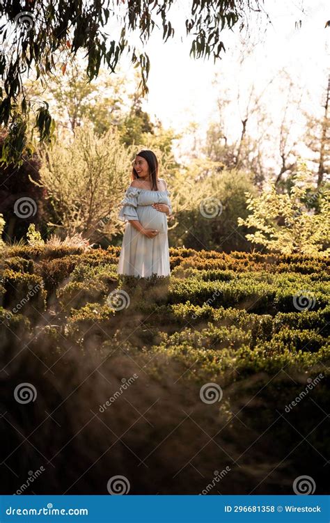 Pregnant Latina Woman In A Dress Standing In Lush Green Sunlit Park Hands Gently Cradling Her