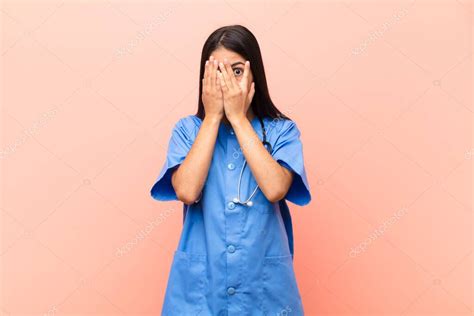 joven enfermera latina cubriendo la cara con las manos asomándose entre los dedos con expresión