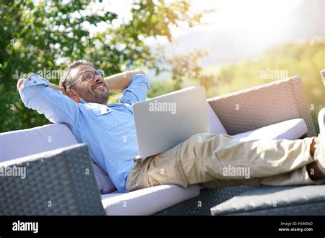Smiling Mature Man Using Laptop Relaxing In Outdoor Sofa Stock Photo