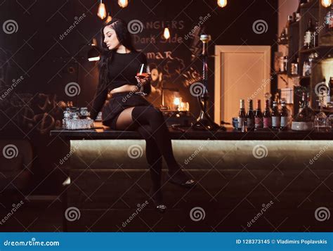 Hot Brunette Girl In Seductive Black Clothes Holds Wineglass While Sitting On The Counter In A