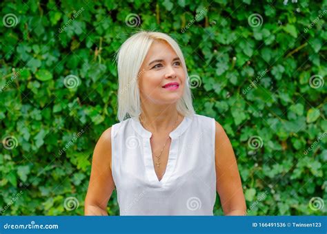 Schöne Blonde Frau Mittleren Alters Im Hintergrund Der Grünen Blätter in Weißer Bluse Auf Der