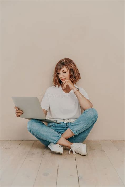 A Brunette Sits On The Floor With A Laptop On A Beige Wall Background She Is Wearing A White T