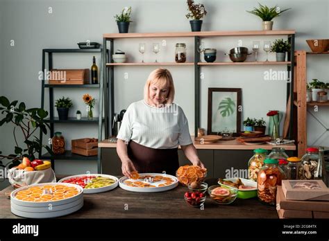 Blond Mature Woman Standing At Counter In Cozy Kitchen And Spreading Fruit Slices On Plastic