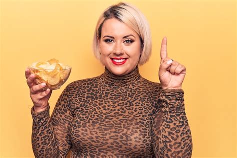 Schöne blonde Plus Size Frau hält eine Schüssel mit Kartoffelchips vor gelbem Hintergrund und