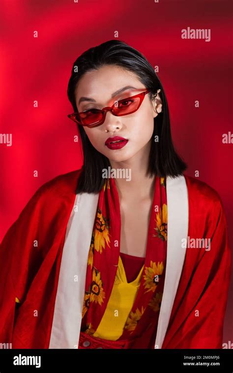 Brunette Asian Woman In Elegant Kimono Cape And Stylish Sunglasses Looking At Camera On Red