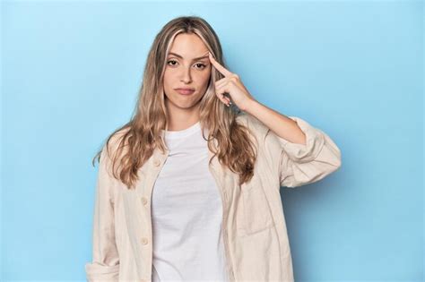 Blonde junge kaukasische Frau im blauen Studio zeigt mit dem Finger auf Schläfe und denkt