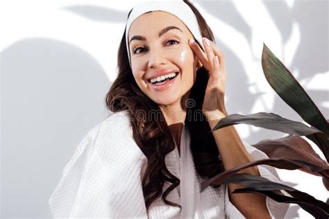 Happy Brunette Woman In A White Bathrobe Takes Care Of Her Skin And Moisturizes Her Face With A