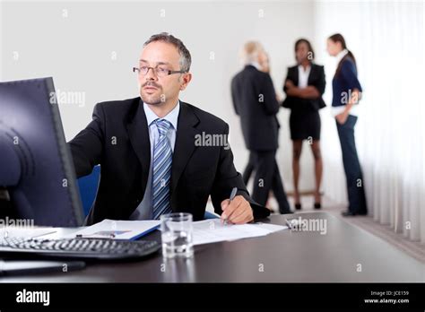 Handsome Mature Business Man Writing Notes While Using Computer At Office With Colleagues Stock