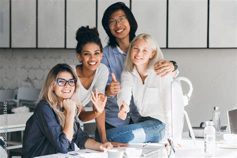 Blissful Asian Programmer Embracing Female Blonde Colleague For Photo And Smiling Indoor