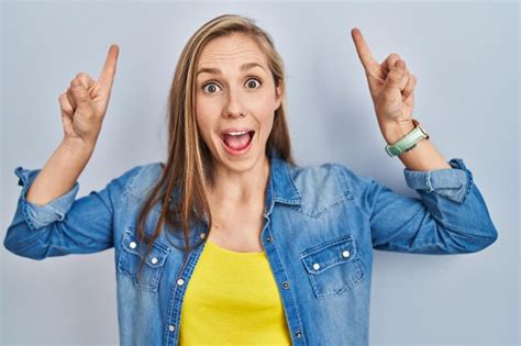 Junge blonde Frau steht vor blauem Hintergrund lächelt erstaunt und überrascht und zeigt mit