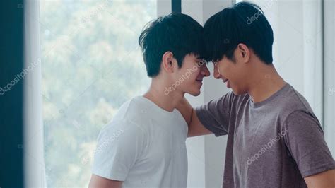 Pareja gay asiática de pie y abrazándose cerca de la ventana en casa Jóvenes asiáticos LGBTQ
