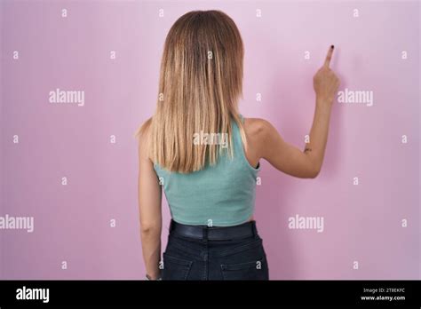 Blonde Caucasian Woman Standing Over Pink Background Posing Backwards Pointing Ahead With Finger