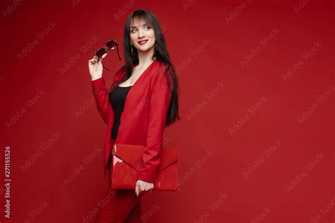 Side View Of Glamorous Brunette Posing In Red Smart Suit And Heels On Red Isolated Background In