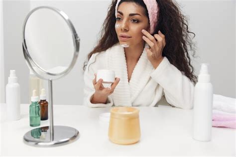 Alegre dama latina rizada y bronceada aplica crema facial en las mejillas admirándose a sí misma