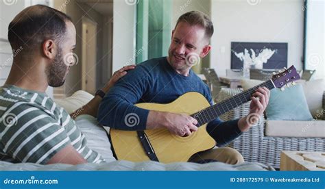 Multi Ethnic Gay Male Couple Sitting On Couch One Playing Guitar Stock