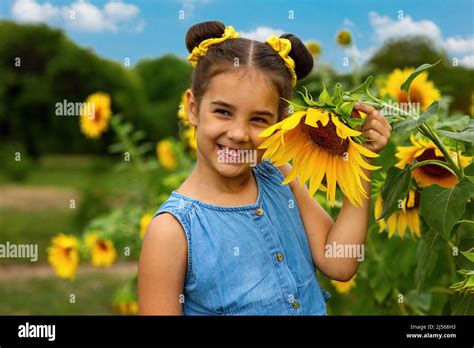 A Happy Little Girl In A Blue Dress Stands Next To The Flowers Of A