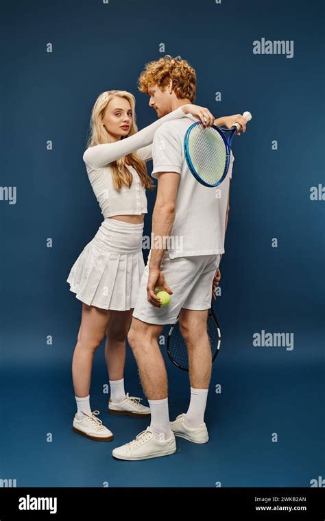 Stylish Blonde Woman With Tennis Racquet Embracing Redhead Man In White Attire On Blue Full