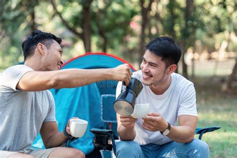Gay LGBTQIA Couple Sitting On Picnic Chair Drinking Tea And Coffee While Camping On Vacation