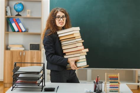 Напряженная молодая учительница, стоящая перед доской и держащая книги ...