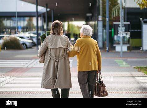 Rear View Of Mature Granddaughter Walking Arm In Arm From Shop Caregiver Carrying Groceries To