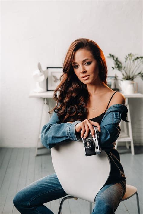 A Brunette With Long Hair Poses In A Studio Sitting On A Chair With A Camera In Her Hands Stock