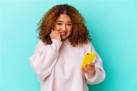 Mujer Latina Joven Que Sostiene El Tel Fono M Vil Aislado En El Fondo Azul Que Muerde Las U As