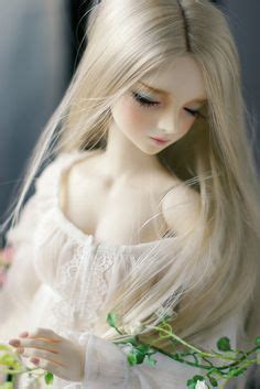 A Doll With Long Blonde Hair Wearing A White Dress And Holding Flowers In Her Hand