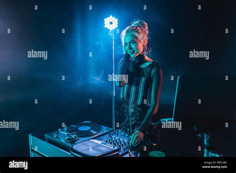 Happy Blonde Dj Girl In Headphones Standing Near Dj Mixer In Nightclub With Smoke Stock Photo