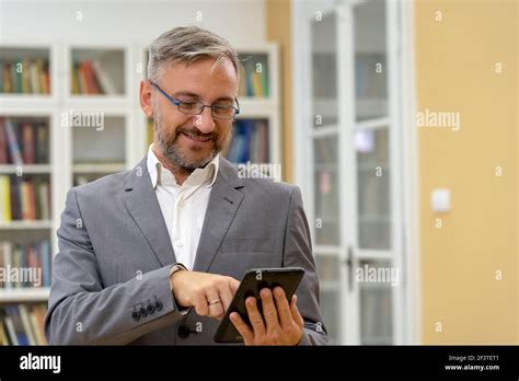 Handsome Mature Businessman Or Teacher Standing In Front Of Bookshelves And Holding Tablet