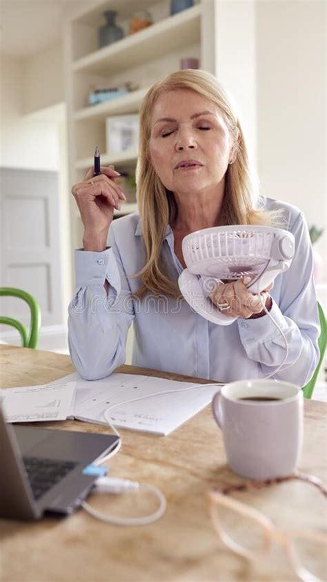 Menopausal Mature Woman Having Hot Flush At Home Cooling Herself With Fan Connected To Laptop