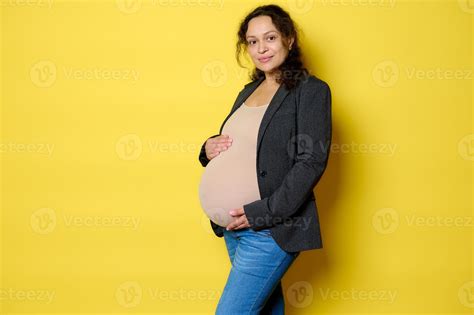 Attractive Latina Pregnant Woman In Stylish Blazer Bodysuit And Blue Jeans Smiles At Camera