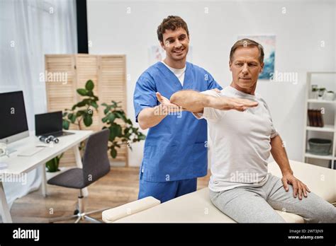 Cheerful Good Looking Doctor In Blue Robe Helping Mature Patient To Stretch During