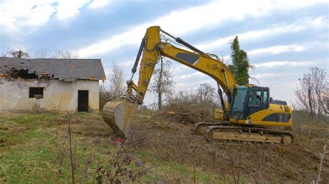 Yellow Excavator Clears Branches Off A Meadow Near The Ruins Of An Old