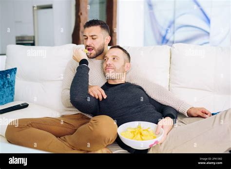 Gay Couple Sitting On The Couch At Home Watching Something On TV And Having A Snack Homosexual
