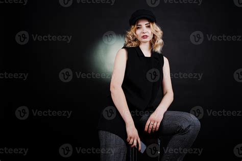 Studio Portrait Of Blonde Girl In Black Wear And Cap With Mobile Phone At Hand Against Dark