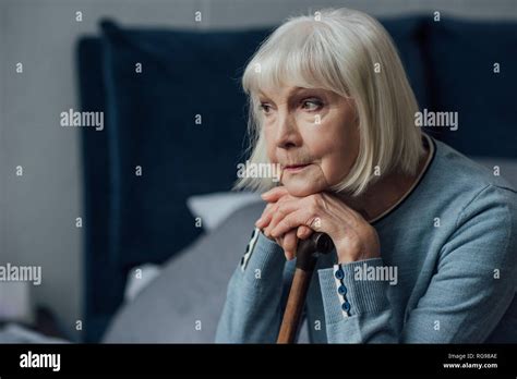 Thoughtful Senior Woman With Hands On Walking Stick Sitting On Bed At