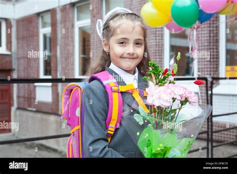 Seven Years Old Girl Standing Behind School Doors With Bag And Flowers