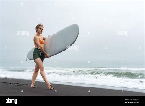 Female Surfer Dressed In Bikini Top Shorts And Barefoot Walks Along Black Sand Beach While
