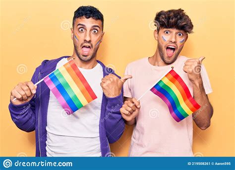 Joven Gay Pareja Sosteniendo Arco Iris Lgbtq Banderas Apuntando Pulgar Hacia El Lado Sonriendo