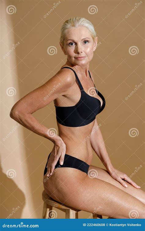 Studio Shot Of Beautiful Mature Woman In Black Underwear Looking At Camera Sitting On A Chair