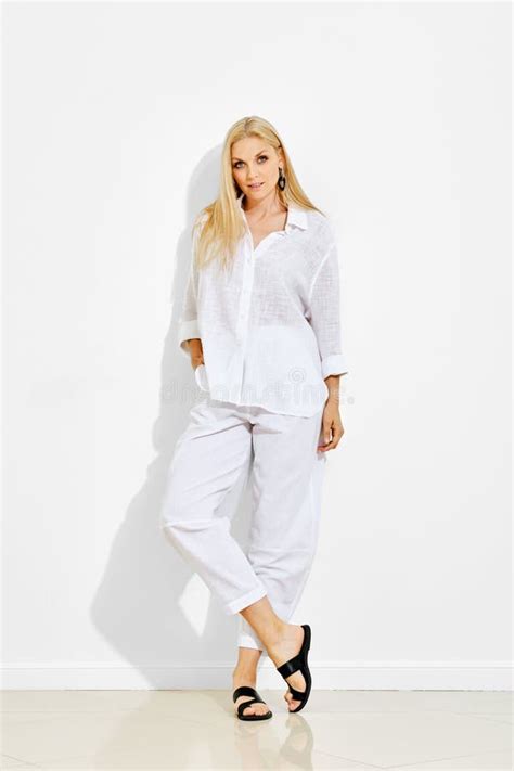 Cute Blonde Woman Standing In Bright Room Against White Wall Wearing Total White Outfit Stock