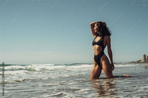 Beautiful Brunette Girl In A Black Swimsuit On The Seashore Stands In The Water Stock Photo