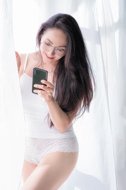 Hermosa mujer latina en lencería blanca parada junto a una ventana y sosteniendo su celular en